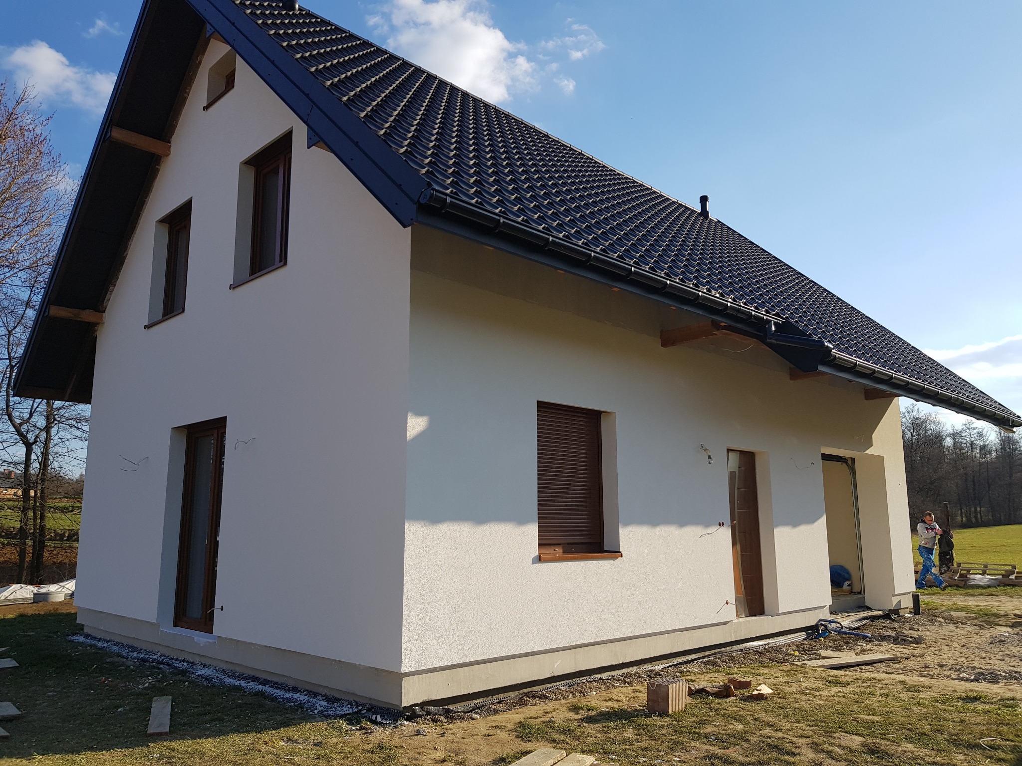 Nowy dom jednorodzinny z jasną elewacją, brązowymi oknami z roletami i ciemną dachówką, widok z boku na tle błękitnego nieba i zielonego krajobrazu, w trakcie prac wykończeniowych.