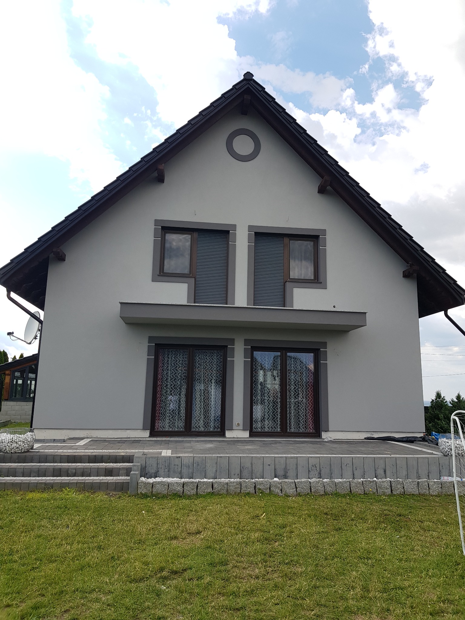 Szary dom jednorodzinny z ciemnobrązowym dachem i stolarką okienną, widok z poziomu trawnika, z elementami dekoracyjnymi z kamienia na schodach i obrzeżach.