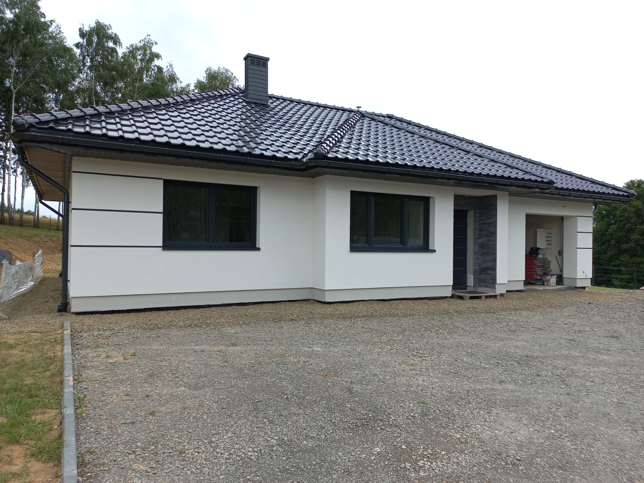 Jednopiętrowy dom z białą elewacją, czarną dachówką i grafitowymi oknami, widok z zewnątrz na podjazd z kamyczków.