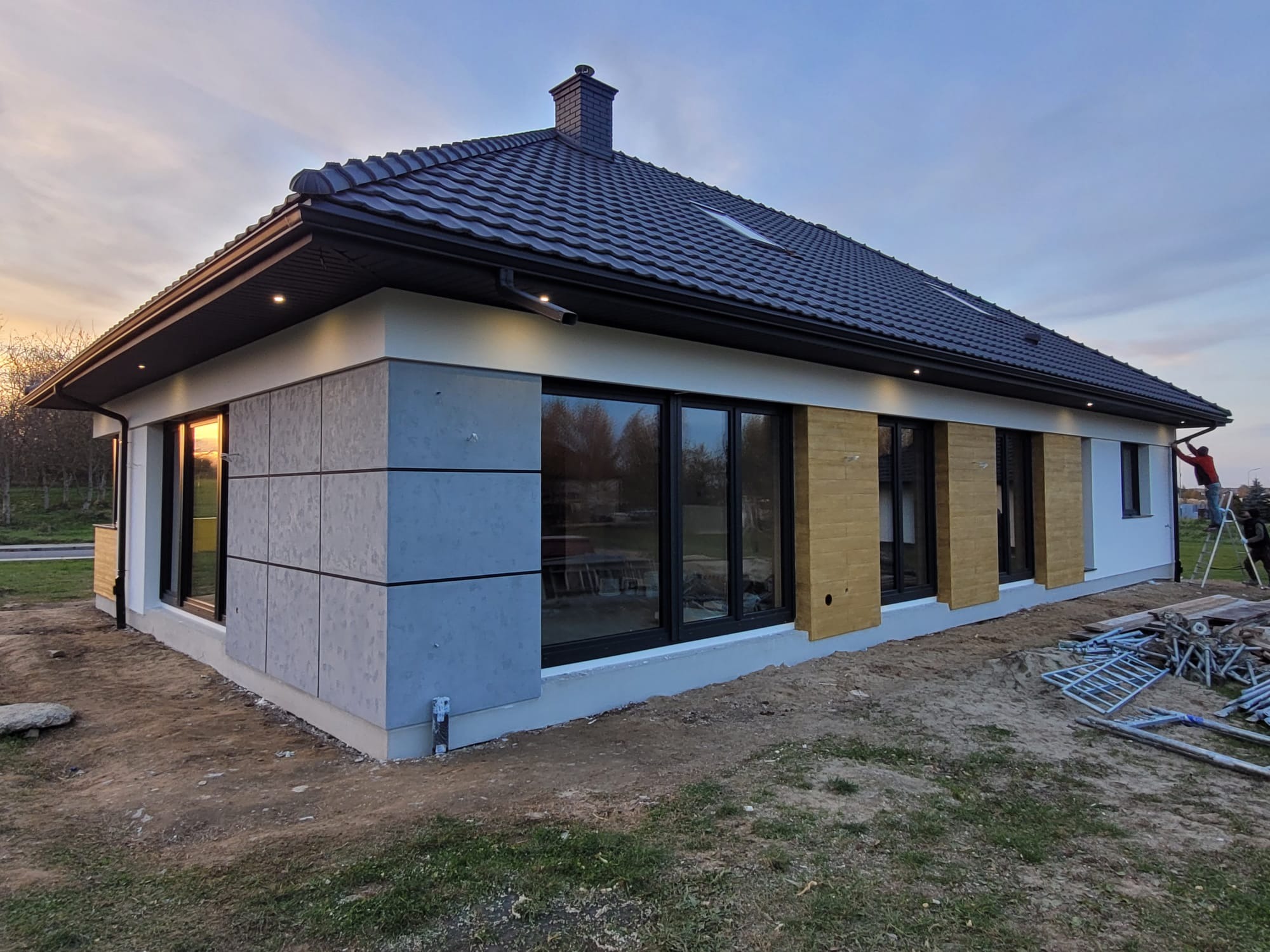 Nowoczesny dom jednorodzinny z elewacją wykończoną panelami w odcieniach szarości i drewna, czarną dachówką i oknami, oświetleniem zewnętrznym i rusztowaniem w tle.