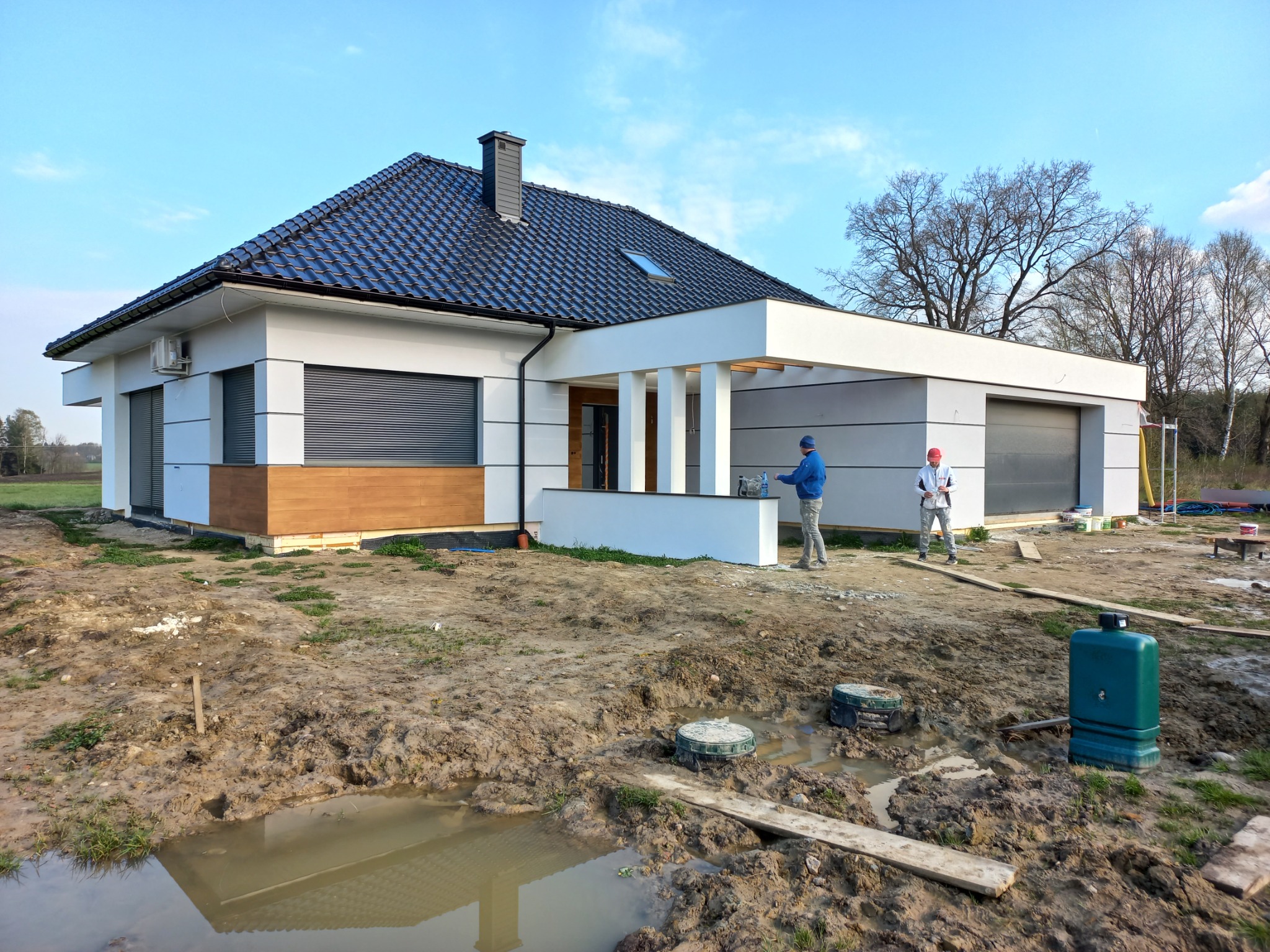 Nowoczesny dom jednorodzinny z szarą dachówką i elementami drewnianej elewacji, widoczni dwaj pracownicy w trakcie wykonywania prac wykończeniowych elewacji, teren wokół domu częściowo rozkopany...