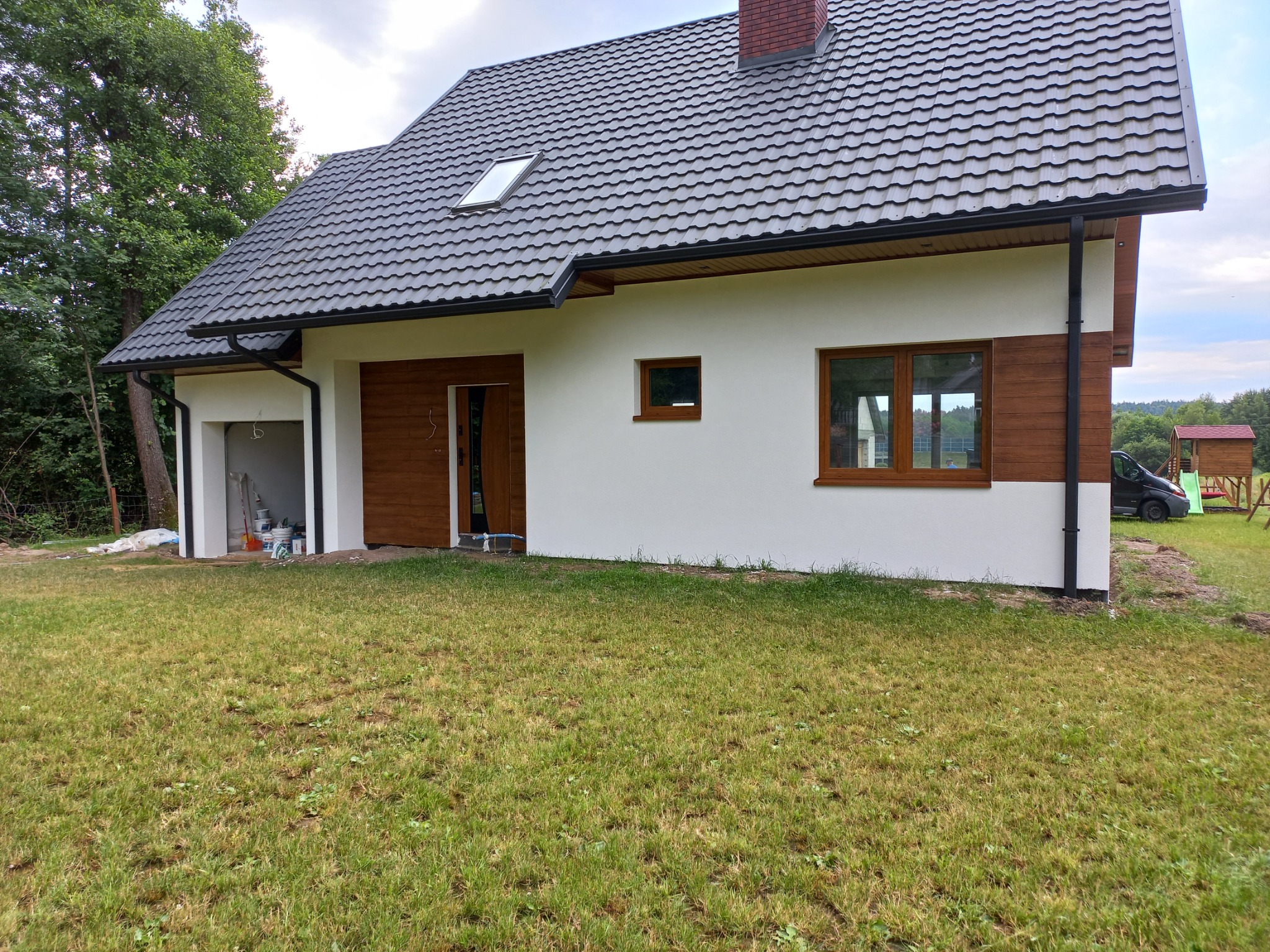 Nowoczesny dom jednorodzinny z białymi ścianami i drewnianymi akcentami, widoczny z perspektywy trawnika, z garażem wbudowanym po lewej stronie i niedokończonym wykończeniem wokół niego.