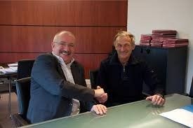 Dwóch mężczyzn w Lisi Ogon uśmiechniętych i podających sobie dłoń nad biurkiem, w tle widoczna szafka z dokumentami i drewniane panele ścienne.