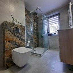 Artdekor - Nowoczesna łazienka z kabiną prysznicową typu walk-in, toaleta podwieszana z dekoracyjnym panelem ściennym imitującym kamień, szare płytki podłogowe i ścienne, oraz szafka umywalkowa z jasnego drewna.