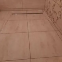 Beżowe płytki podłogowe z odpływem liniowym w narożniku łazienki, wzorzysta tapeta w tle.