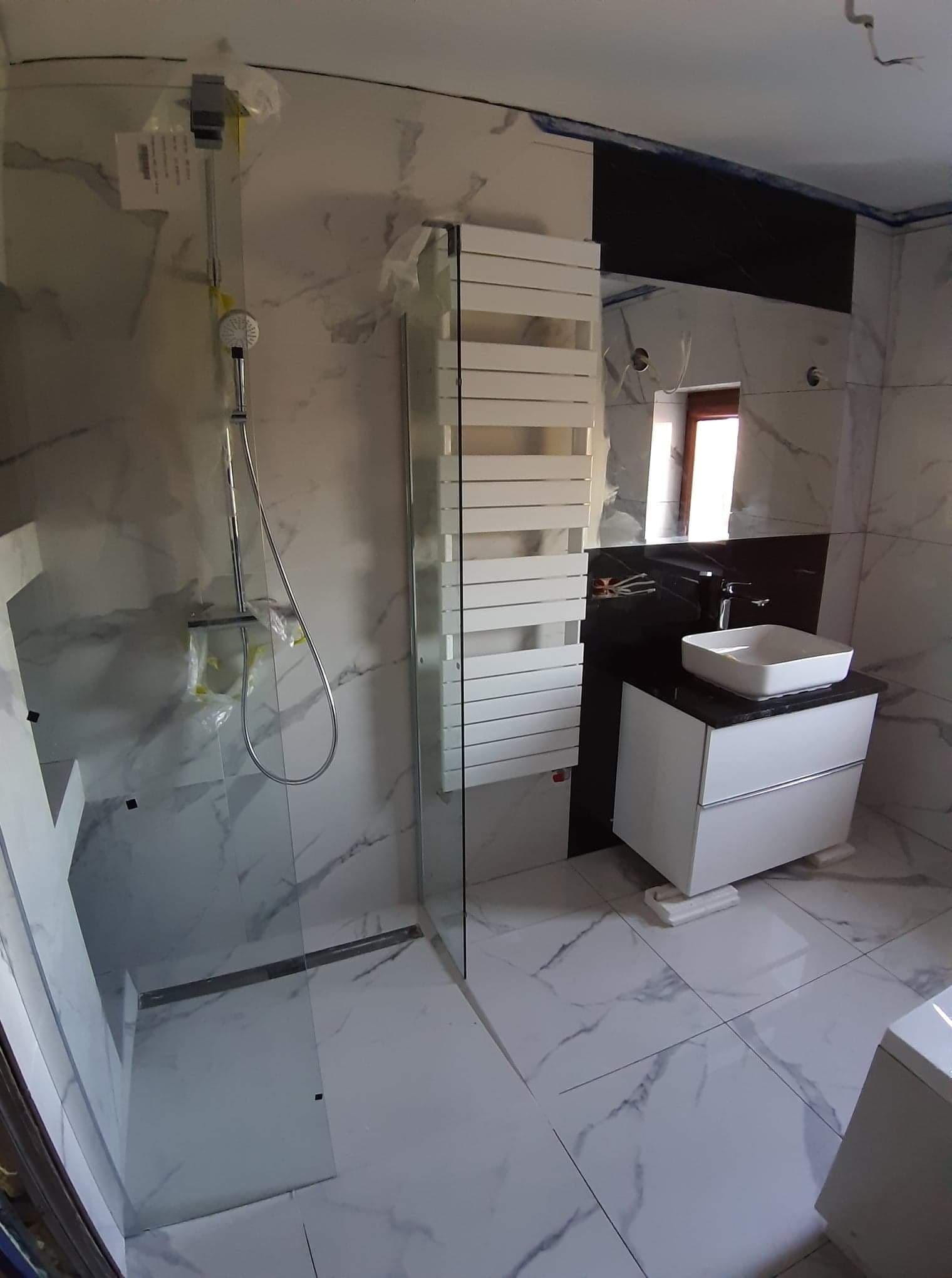Nowoczesna łazienka w trakcie wykańczania: białe marmurowe płytki, szklana kabina prysznicowa z zabezpieczoną folią słuchawką, biała szafka z umywalką na czarnym blacie i grzejnik drabinkowy.