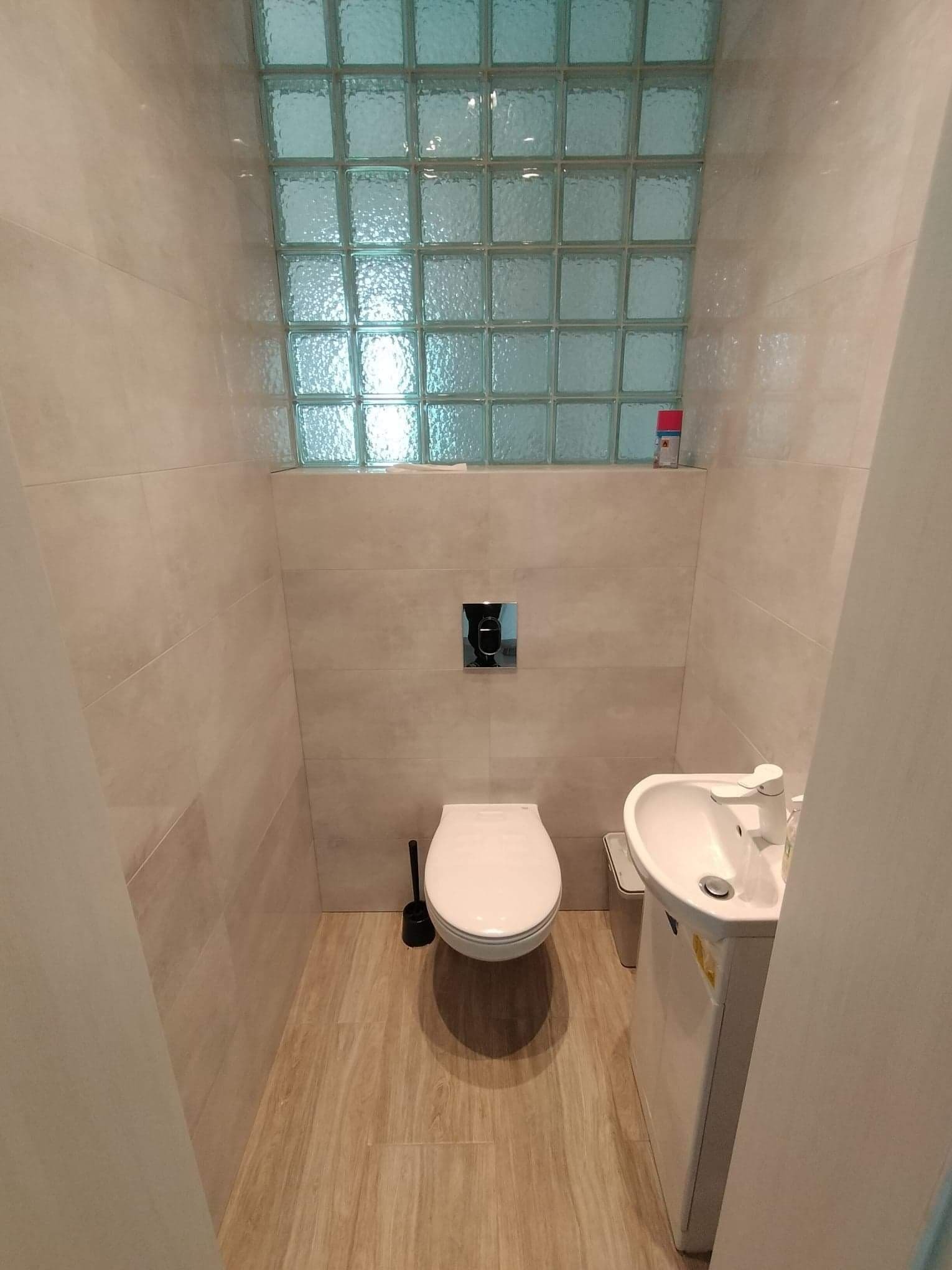 Mała łazienka z beżowymi płytkami, panelem z luksferów, wiszącą toaletą i umywalką z szafką, na podłodze panele imitujące drewno.