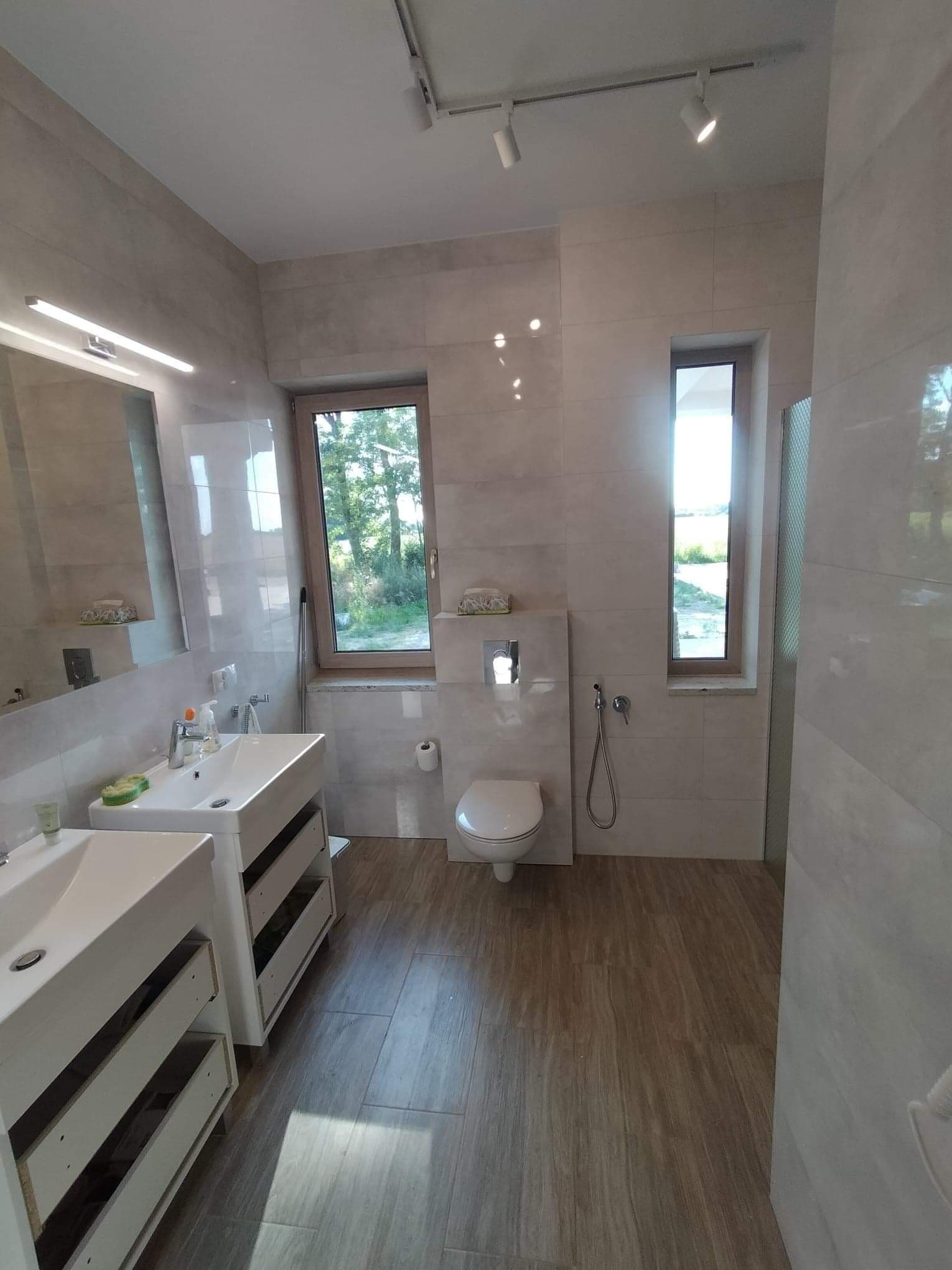 Jasna łazienka po remoncie z dwoma umywalkami na szafkach z otwartymi szufladami, toaleta pod oknem i płytki imitujące drewno na podłodze.