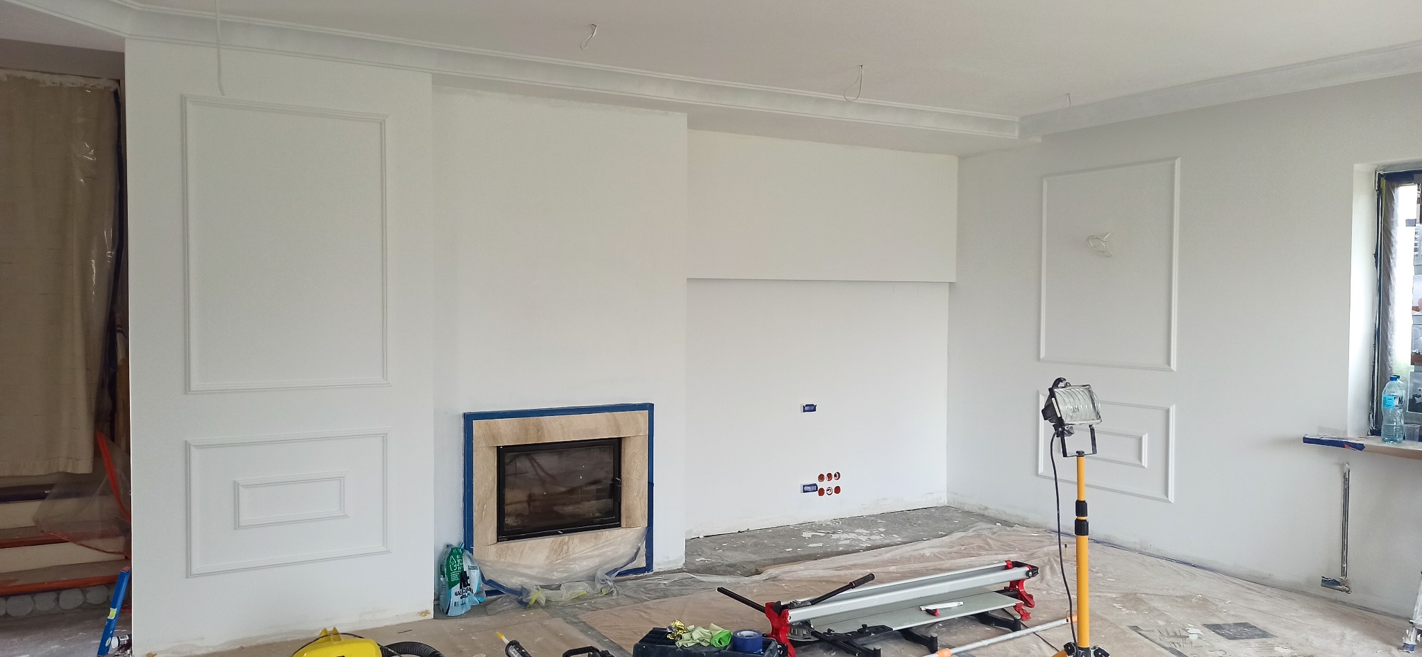 Pokój w trakcie remontu: świeżo pomalowane na biało ściany z dekoracyjnymi sztukateriami, obudowa kominka z beżowego kamienia zabezpieczona folią, widoczne instalacje elektryczne, narzędzia...