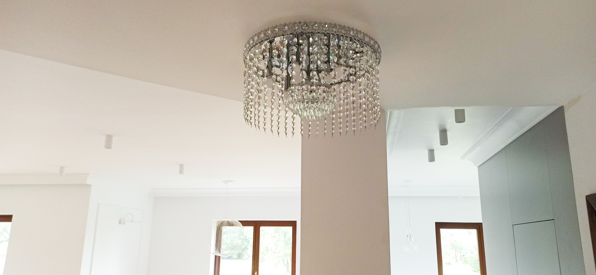 Kryształowy żyrandol sufitowy w jasnym wnętrzu, widoczne listwy dekoracyjne i minimalistyczne lampy punktowe