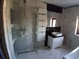 Wnętrze łazienki w trakcie prac wykończeniowych: marmurowe płytki na ścianach i podłodze, zamontowana kabina prysznicowa z folią ochronną, biały grzejnik drabinkowy i umywalka nablatowa na szafce...