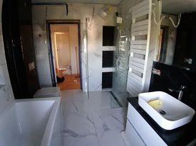 Wnętrze łazienki w trakcie remontu: białe marmurowe płytki na podłodze i ścianach, widoczna wanna, umywalka nablatowa, grzejnik drabinkowy zabezpieczony folią, otwarte drzwi do dalszej części...