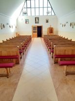 Wnętrze kaplicy z jasnymi płytkami podłogowymi, rzędami drewnianych ławek z czerwonymi siedziskami i wysokim oknem w kształcie trójkąta, wpadające światło słoneczne.