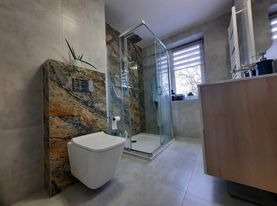 Nowoczesna łazienka z kabiną prysznicową typu walk-in, toaleta podwieszana z dekoracyjnym panelem ściennym imitującym kamień, szare płytki podłogowe i ścienne, oraz szafka umywalkowa z jasnego drewna.