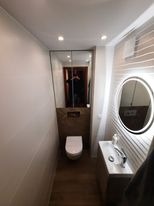 Wąska toaleta w jasnych barwach z umywalką i okrągłym lustrem podświetlanym LED. Na ścianie za toaletą dekoracyjny panel imitujący kamień. Widoczne oświetlenie punktowe w suficie.