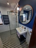 Nowoczesna łazienka z czarno-białą mozaiką na podłodze, białe kafelki imitujące cegłę na ścianach, kabina prysznicowa z czarnymi profilami, owalne lustro w miedzianej ramie na granatowej ścianie...