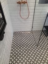 Wykończona kabina prysznicowa z białą, cegiełkową glazurą na ścianach i wzorzystą, geometryczną posadzką w odcieniach szarości i bieli. Miedziana bateria prysznicowa z widocznym wężem. Fragment...