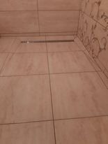 Beżowe płytki podłogowe z odpływem liniowym w narożniku łazienki, wzorzysta tapeta w tle.