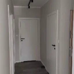 Szary korytarz z ciemną podłogą z płytek, białymi drzwiami z czarnymi klamkami i czarnym oświetleniem szynowym.