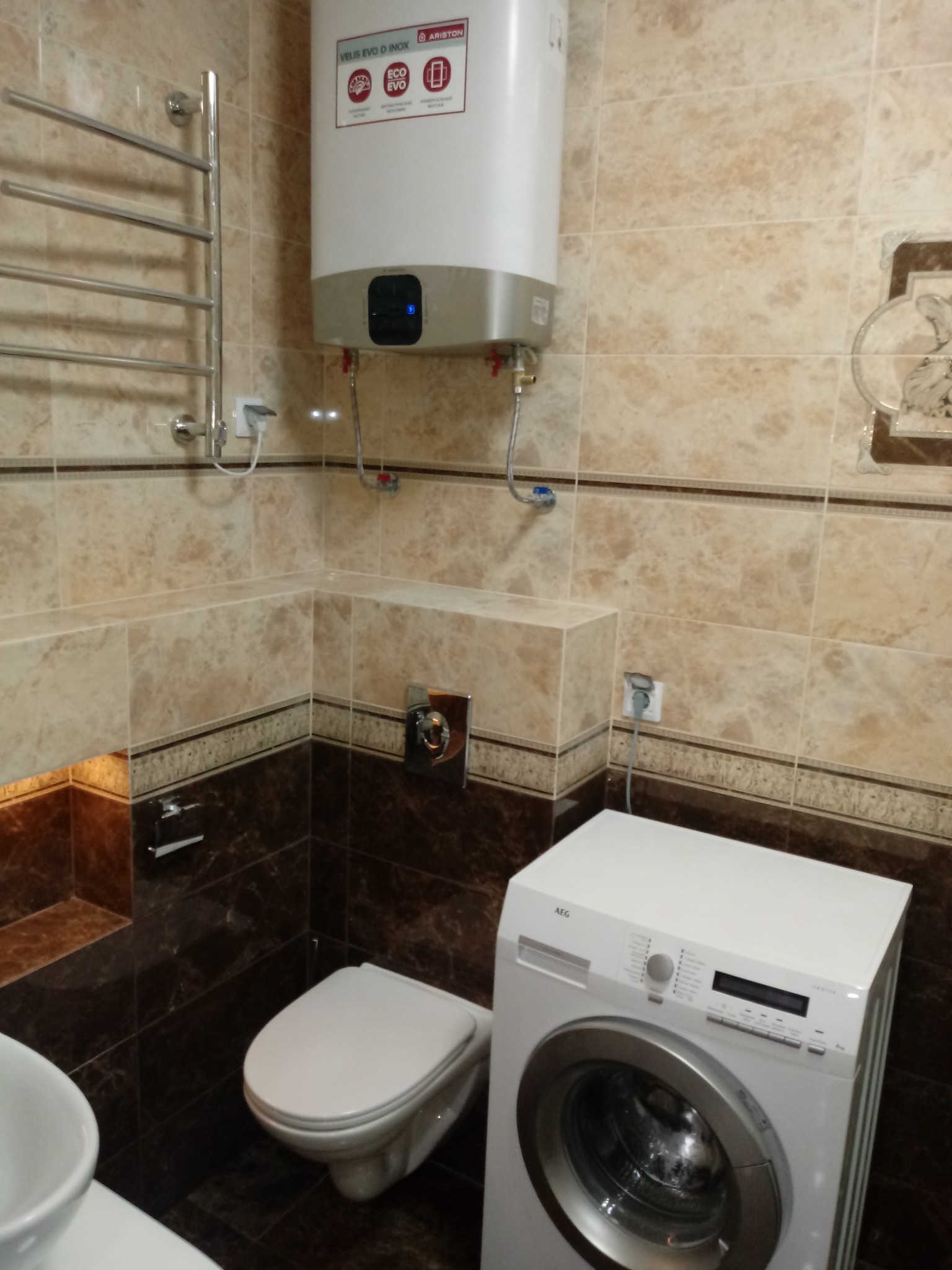 Beżowo-brązowa łazienka z bojlerem Ariston, grzejnikiem drabinkowym, toaletą podwieszaną i pralką AEG.
