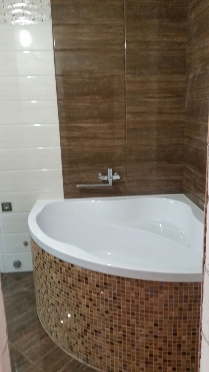 Biała wanna narożna z mozaikową obudową, brązowe płytki imitujące drewno na ścianie, chromowana bateria.