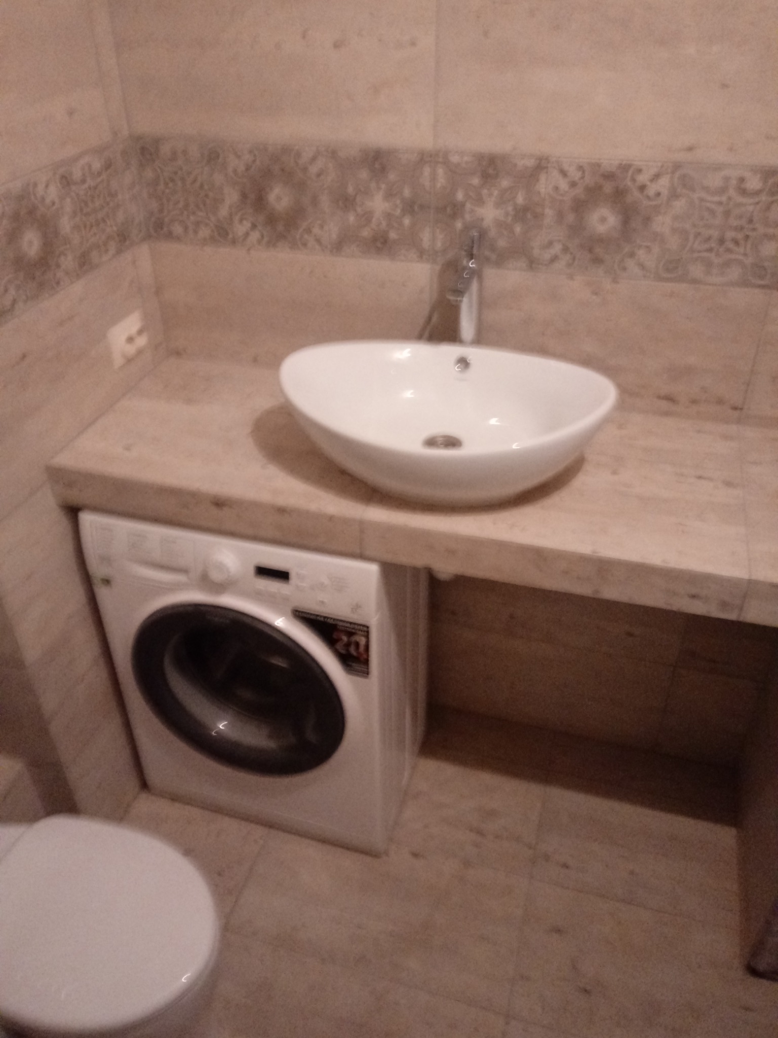 Umywalka nablatowa w kształcie misy z chromowaną baterią, zamontowana na blacie z jasnego kamienia, pod którym znajduje się pralka automatyczna. W tle dekoracyjna listwa z motywem roślinnym...