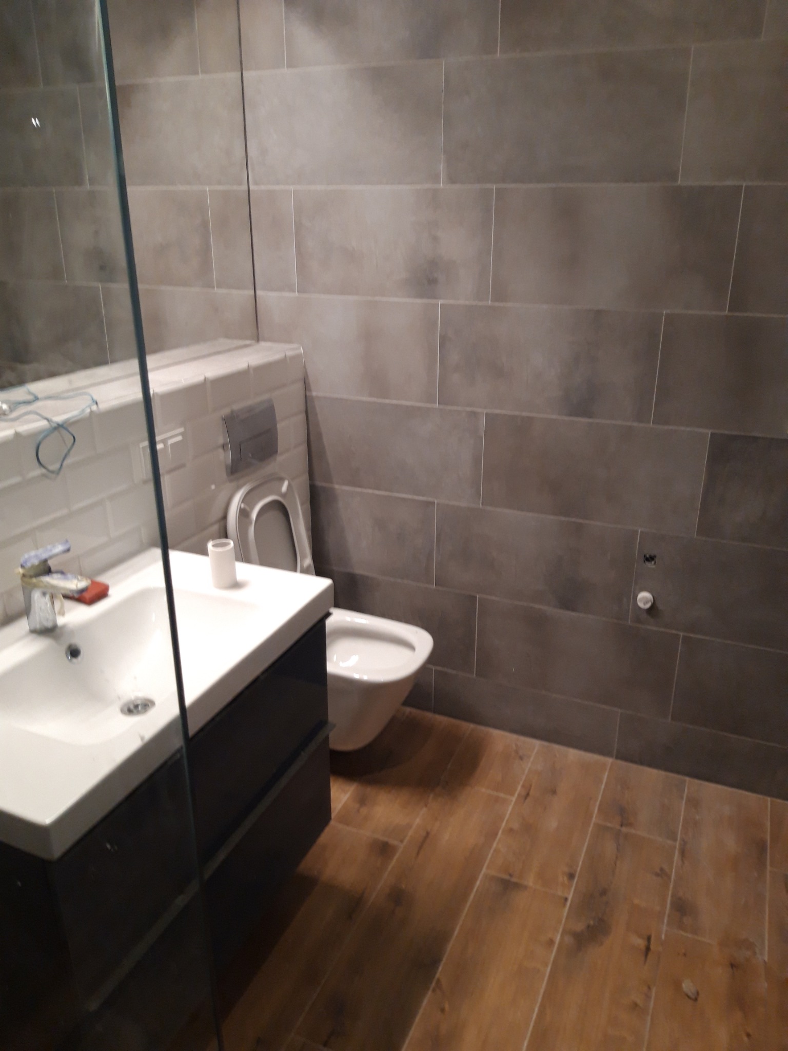 Nowoczesna łazienka z szarymi płytkami imitującymi kamień na ścianach, drewnopodobną podłogą, białą umywalką z szafką oraz wiszącą miską WC, widok zza szklanej kabiny.