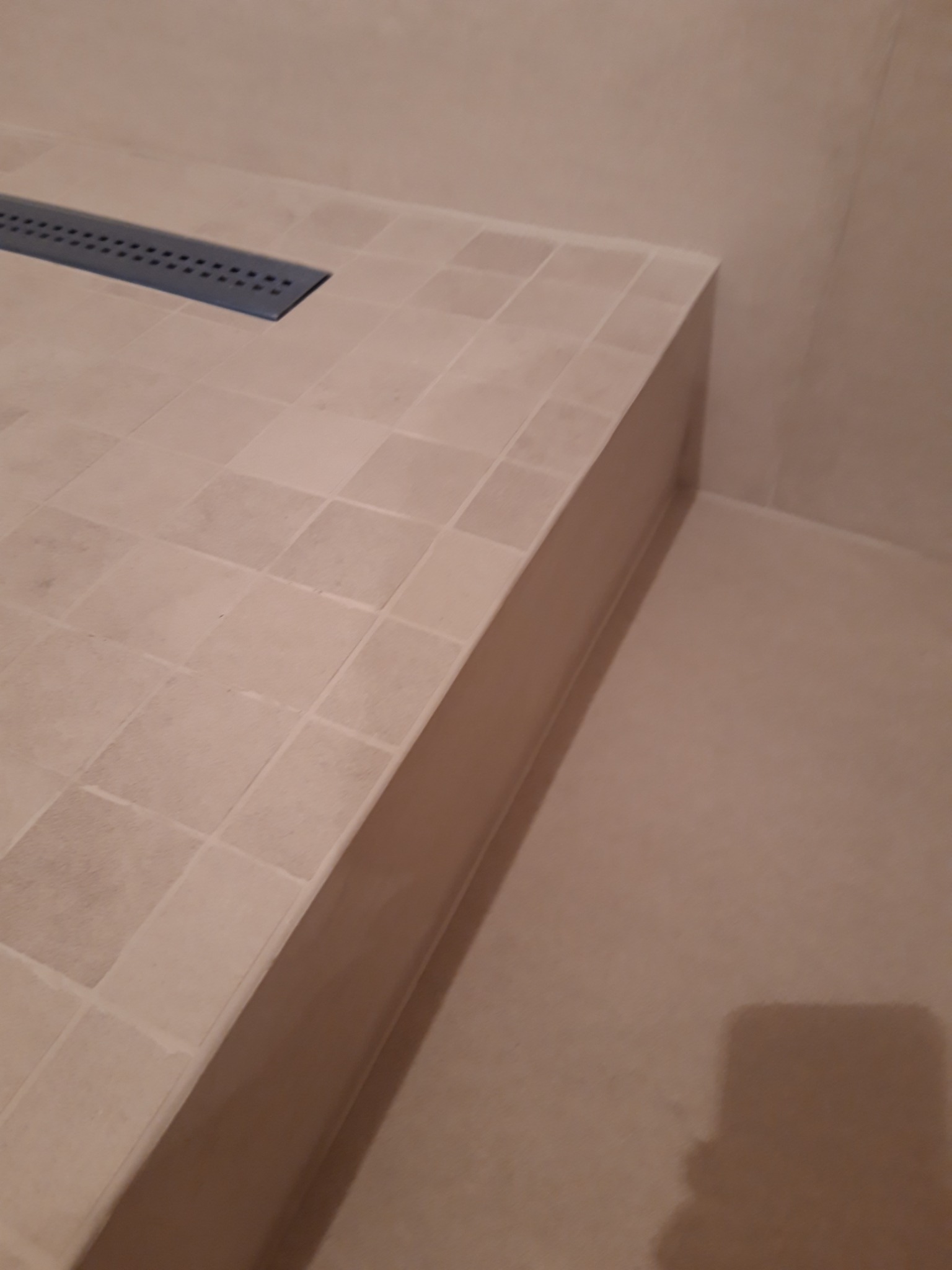 Beżowa, kwadratowa mozaika ceramiczna w strefie prysznicowej z odpływem liniowym w rogu.