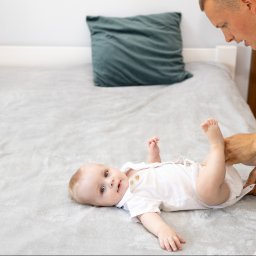 Izabela Marosek Fotografia - Ojciec w białej koszuli zapina bodziaka leżącemu na szarym łóżku niemowlęciu z niebieskimi oczami, które patrzy w obiektyw.