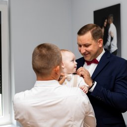 Izabela Marosek Fotografia - Mężczyzna w granatowym garniturze z bordową muchą poprawia muchę niemowlęciu ubranemu w białą koszulę i beżowe szelki, drugi mężczyzna w białej koszuli patrzy na nich. W tle okno i czarno-białe...