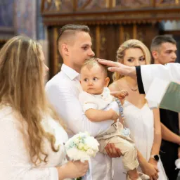 Ksiądz w białej szacie błogosławi niemowlę trzymane przez ojca podczas ceremonii w kościele, obok stoi matka z bukietem białych kwiatów.