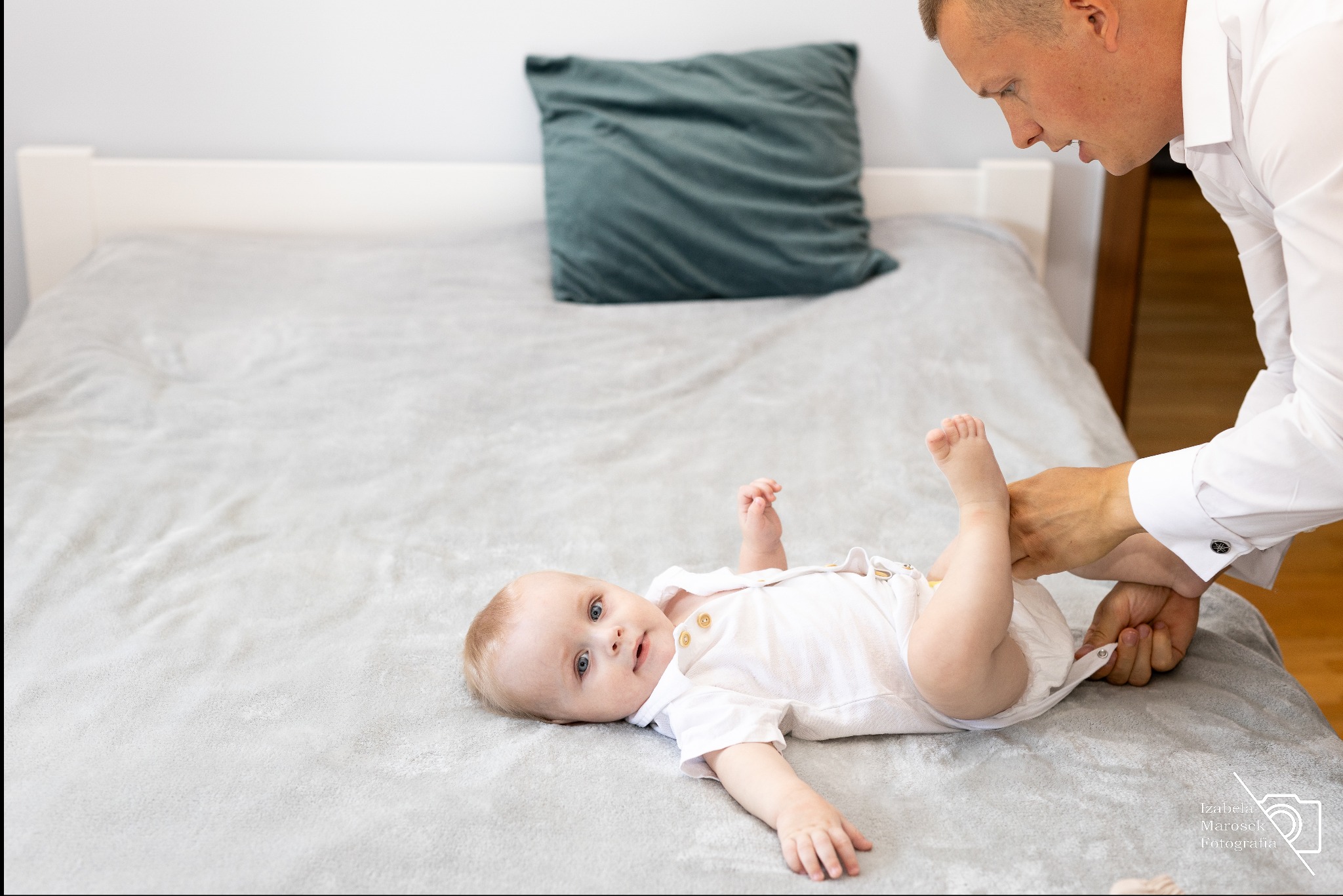 Ojciec w białej koszuli zapina bodziaka leżącemu na szarym łóżku niemowlęciu z niebieskimi oczami, które patrzy w obiektyw.