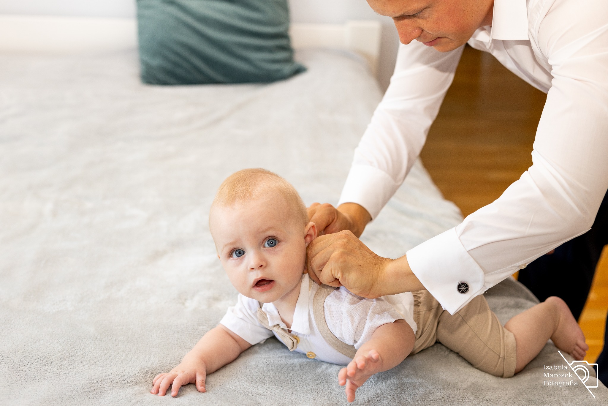 Niemowlę leżące na brzuchu, ubrane w koszulę i beżowe spodnie z szelkami, ojciec poprawia mu ubranie, widoczne mankiety koszuli ojca ze spinkami.