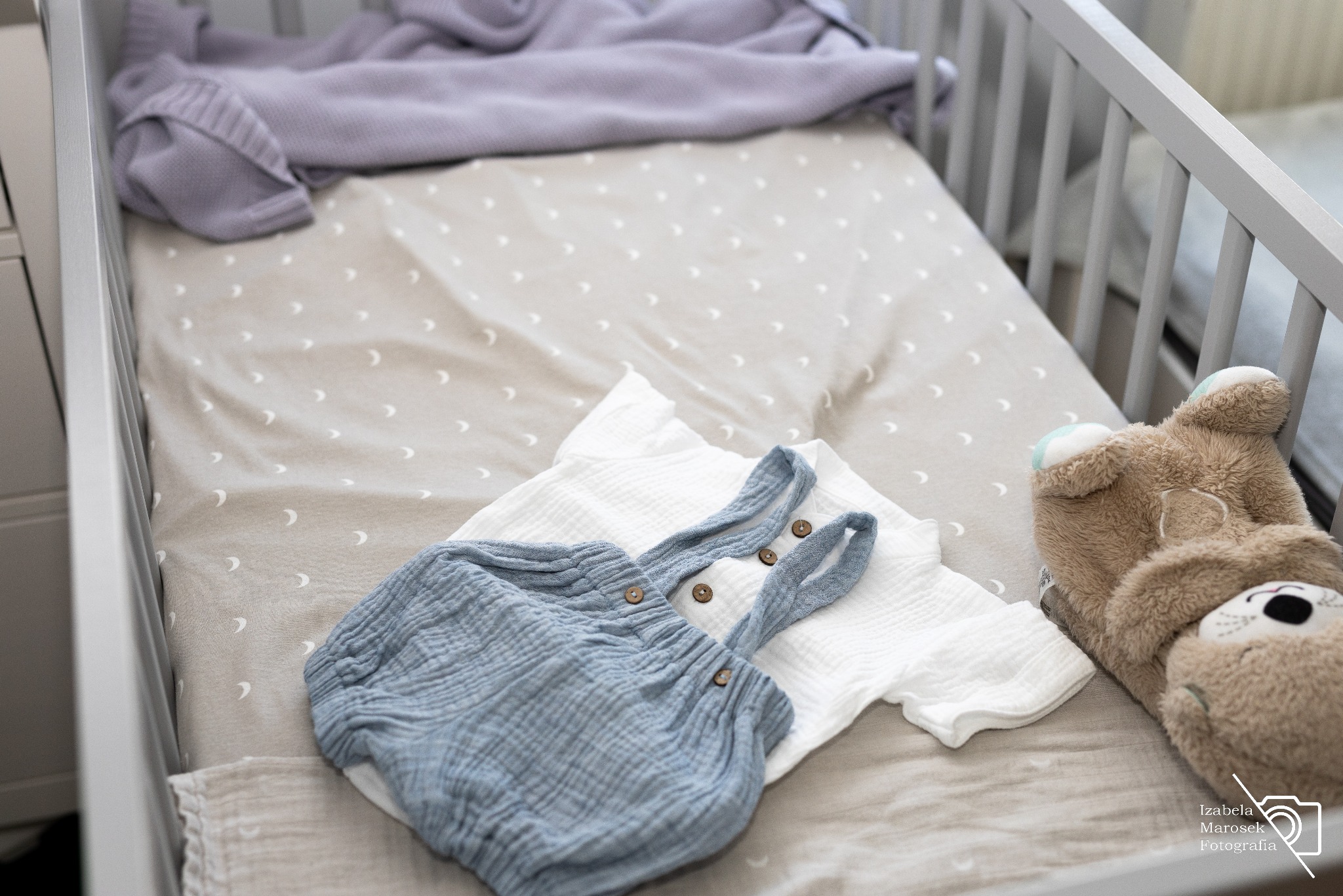 Ubranka niemowlęce z lnu i pluszowy miś w białym łóżeczku z pościelą w księżyce, przygotowane do sesji noworodkowej.