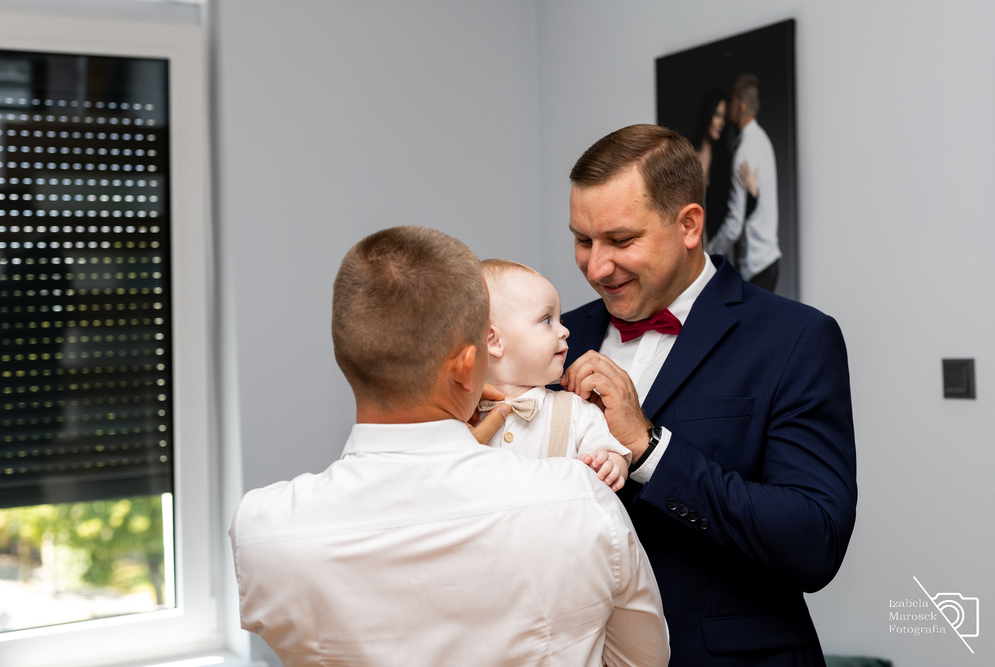 Mężczyzna w granatowym garniturze z bordową muchą poprawia muchę niemowlęciu ubranemu w białą koszulę i beżowe szelki, drugi mężczyzna w białej koszuli patrzy na nich. W tle okno i czarno-białe...