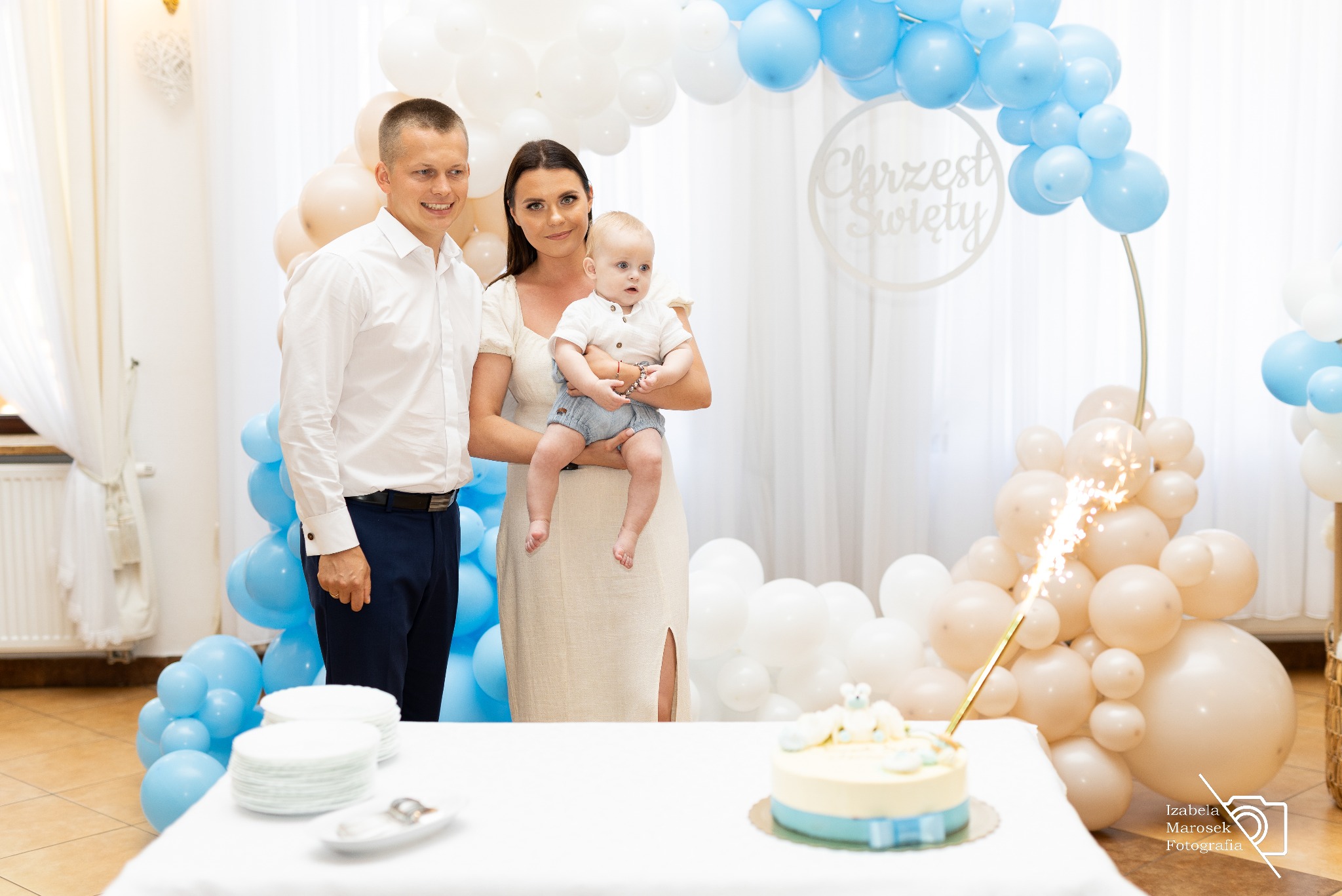 Uśmiechnięta rodzina pozuje na tle dekoracji z balonów w kolorach błękitu i bieli, tort z fontanną iskrzy się na stole podczas uroczystości Chrztu Świętego.