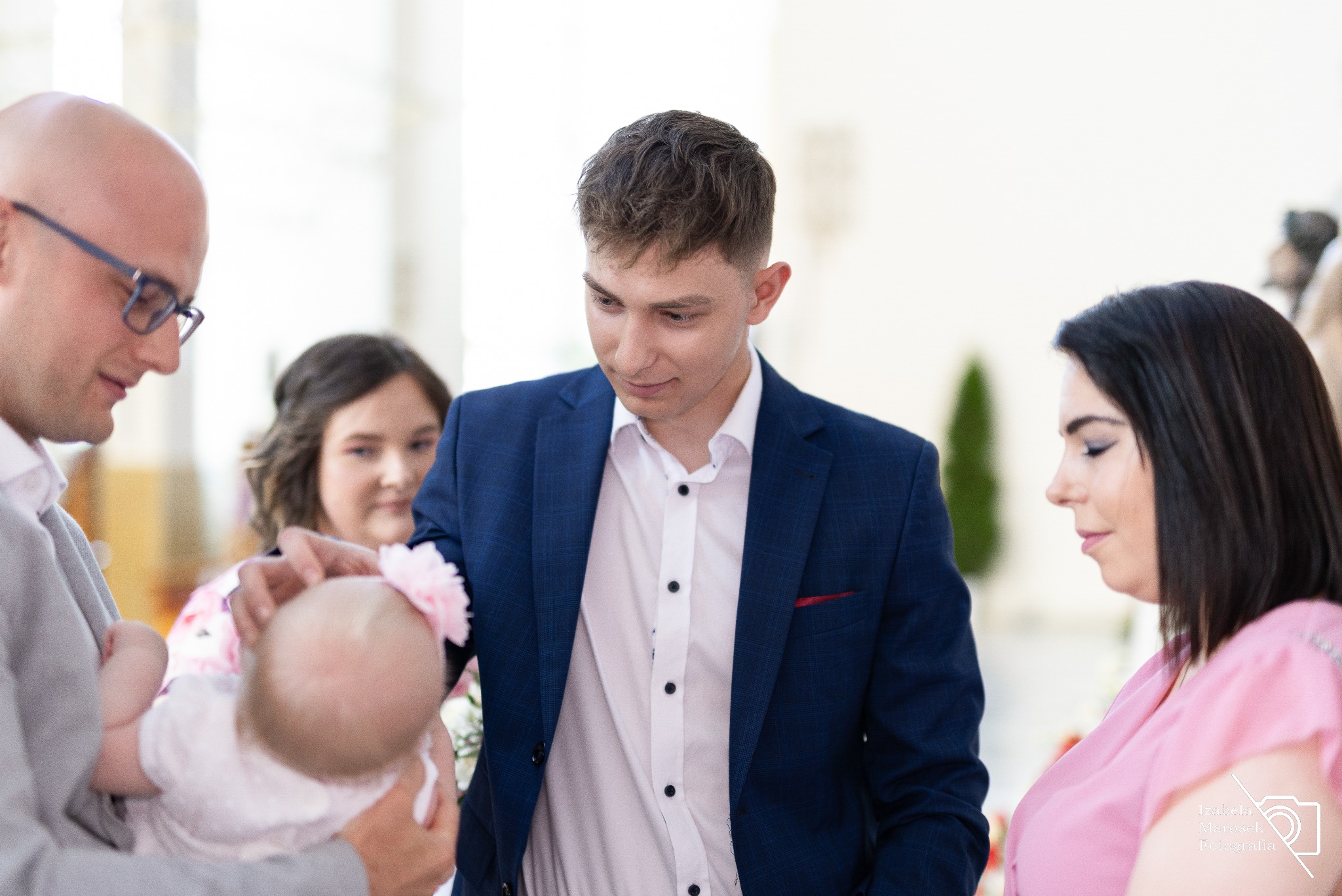 Uroczystość chrztu: mężczyzna w okularach trzyma niemowlę z różową ozdobą we włosach, obok młody mężczyzna w granatowej marynarce i kobieta w różowej sukience, w tle dziewczynka.