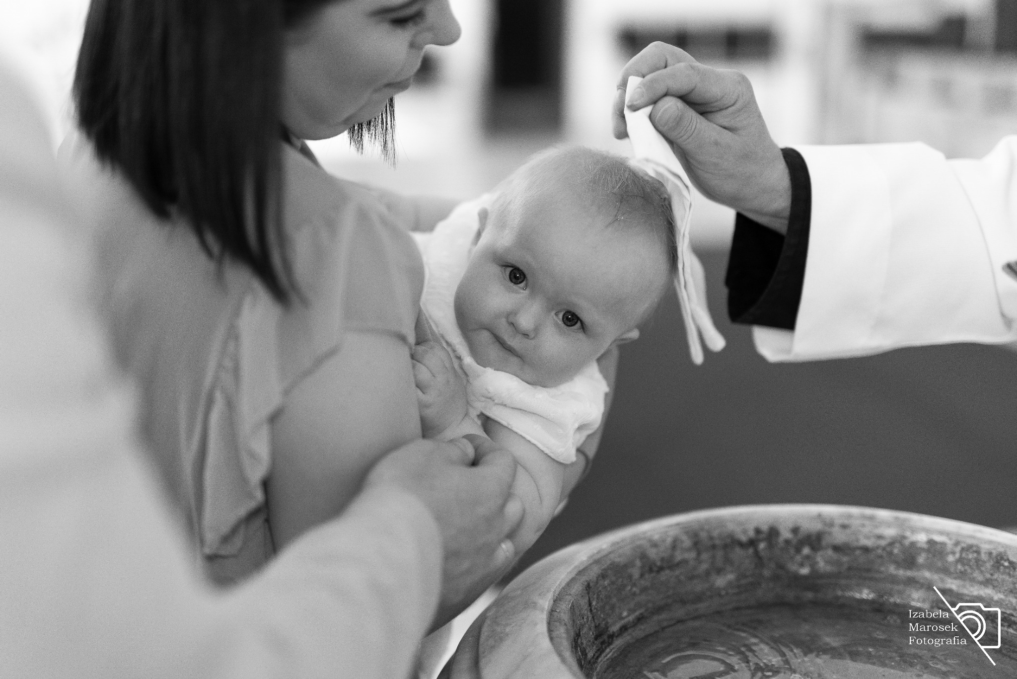 Czarno-białe zdjęcie przedstawia niemowlę w ramionach matki podczas chrztu, z widoczną chrzcielnicą i ręką księdza polewającą wodę na główkę dziecka. Uwagę przykuwa wyraz zaciekawienia na twarzy...