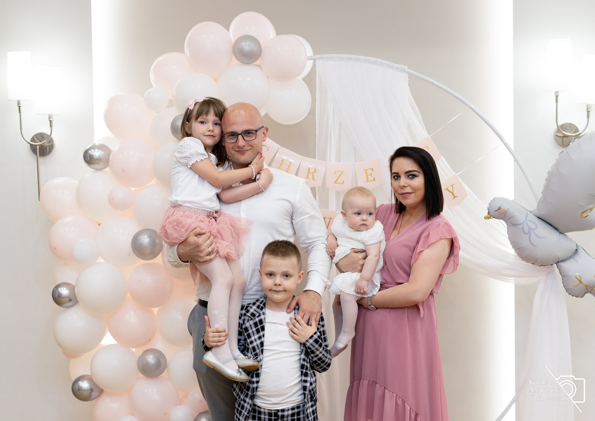 Uśmiechnięta rodzina pozuje na tle dekoracji z balonów i napisu 'Chrzest', mama trzyma niemowlę w białej sukience, tata obejmuje starszą córkę w różowej spódniczce, a syn stoi przed nim w koszuli...
