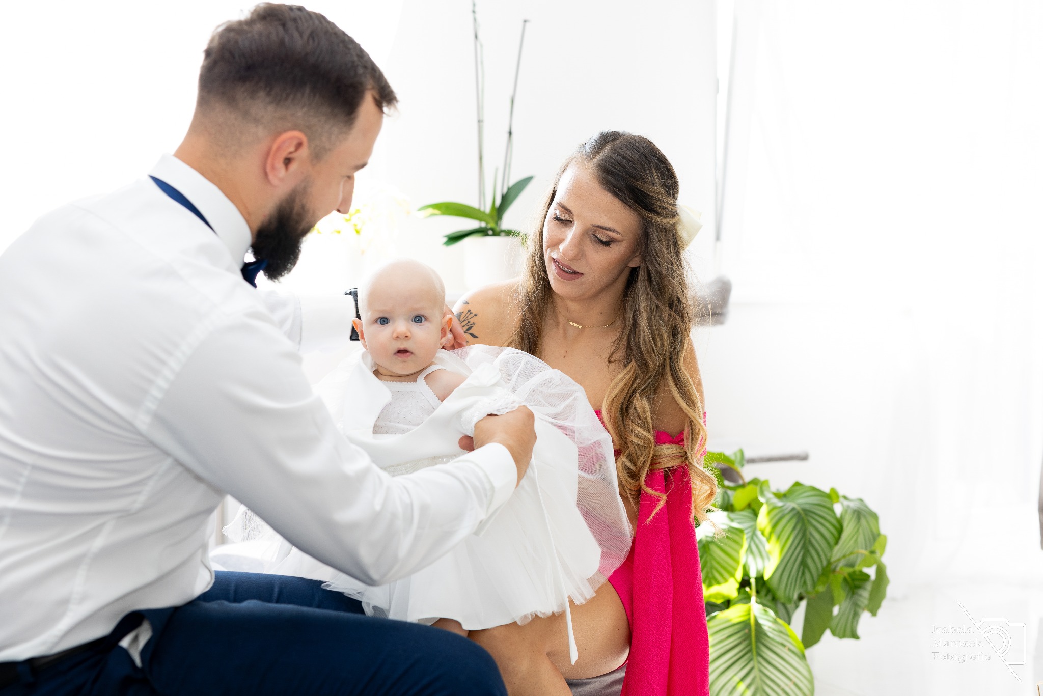 Rodzice ubierają niemowlę w białą sukienkę, przygotowując je do uroczystości, widoczny fragment wnętrza z rośliną w tle.