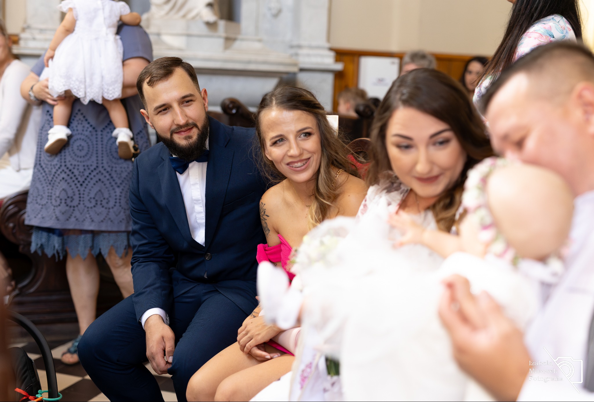 Uśmiechnięci rodzice i goście podczas chrztu dziecka w kościele, widoczna mama z aparatem ortodontycznym i tatuażem na ramieniu, elegancki ojciec w granatowym garniturze i mucha.