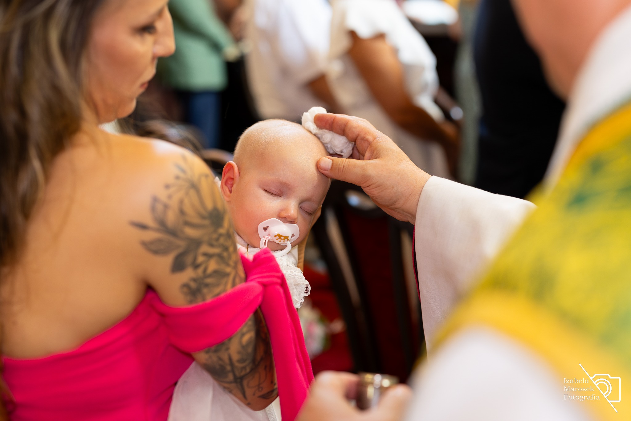 Kadr z ceremonii chrztu: niemowlę z zamkniętymi oczami i smoczkiem, trzymane przez matkę z tatuażem na ramieniu, ksiądz dokonuje namaszczenia czoła watą.