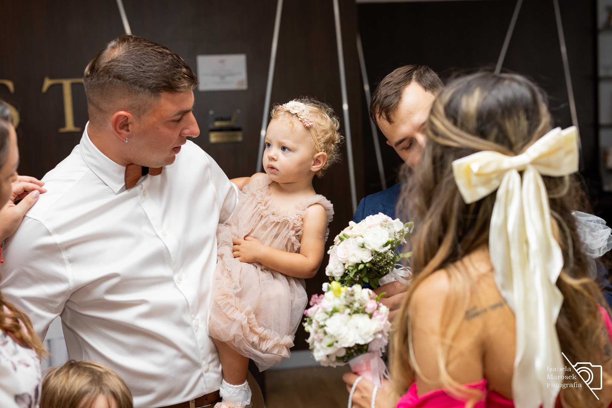 Uroczystość rodzinna: mężczyzna trzyma na rękach małą dziewczynkę w tiulowej sukience, obok stoi osoba z bukietem białych róż, w tle ciemne drewniane panele.