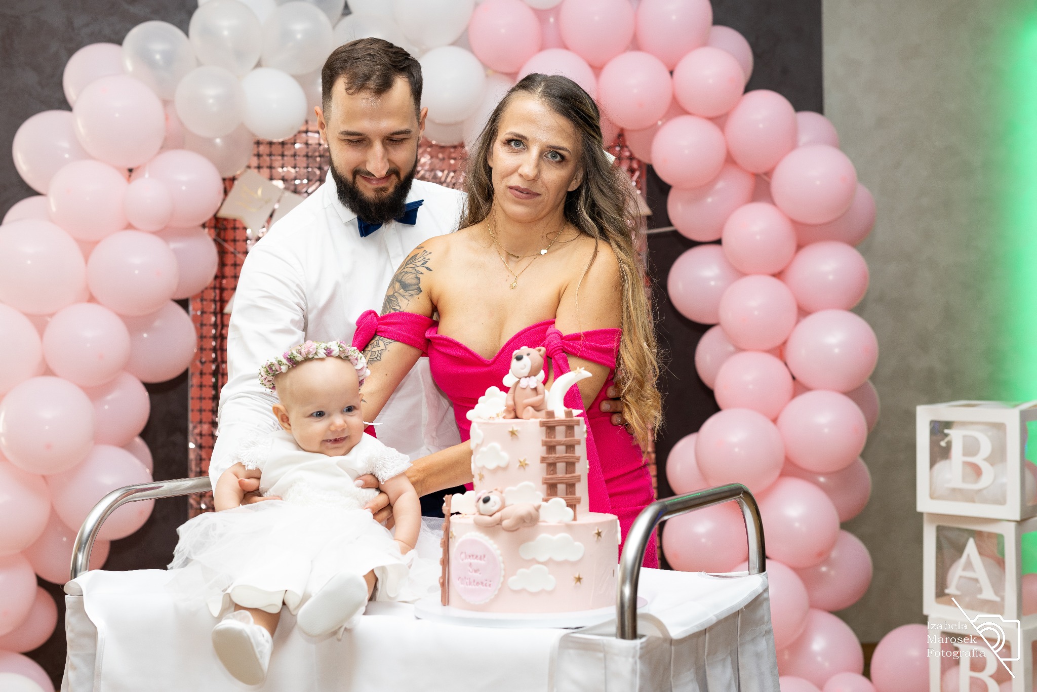 Rodzina pozuje przy torcie z dekoracjami misia i księżyca, otoczona różowymi i białymi balonami oraz dekoracyjnymi kostkami z napisem BABY.