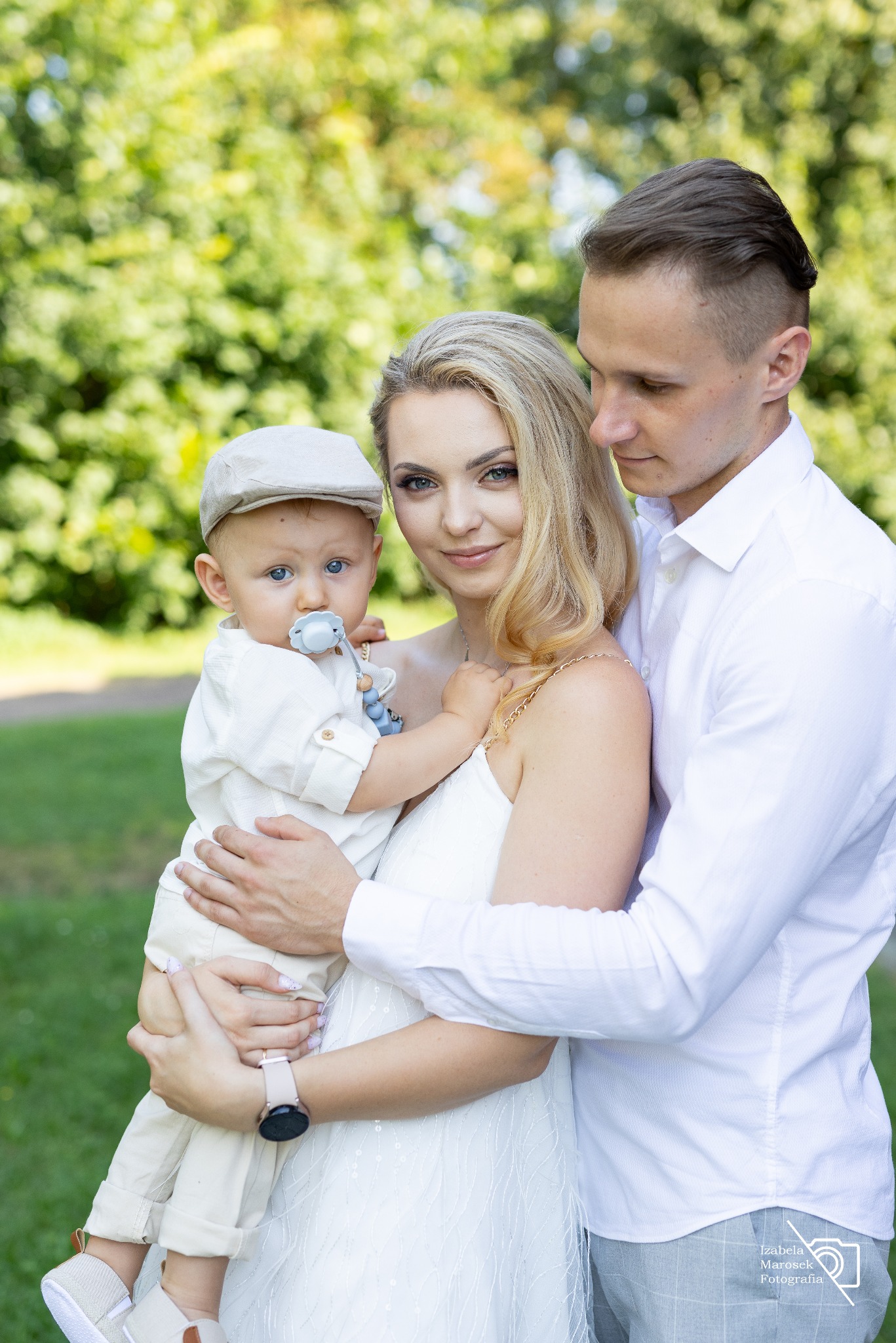 Portret młodej rodziny: matka trzyma na rękach synka w kaszkiecie, ojciec obejmuje żonę, wszyscy ubrani w jasne kolory na tle zieleni.
