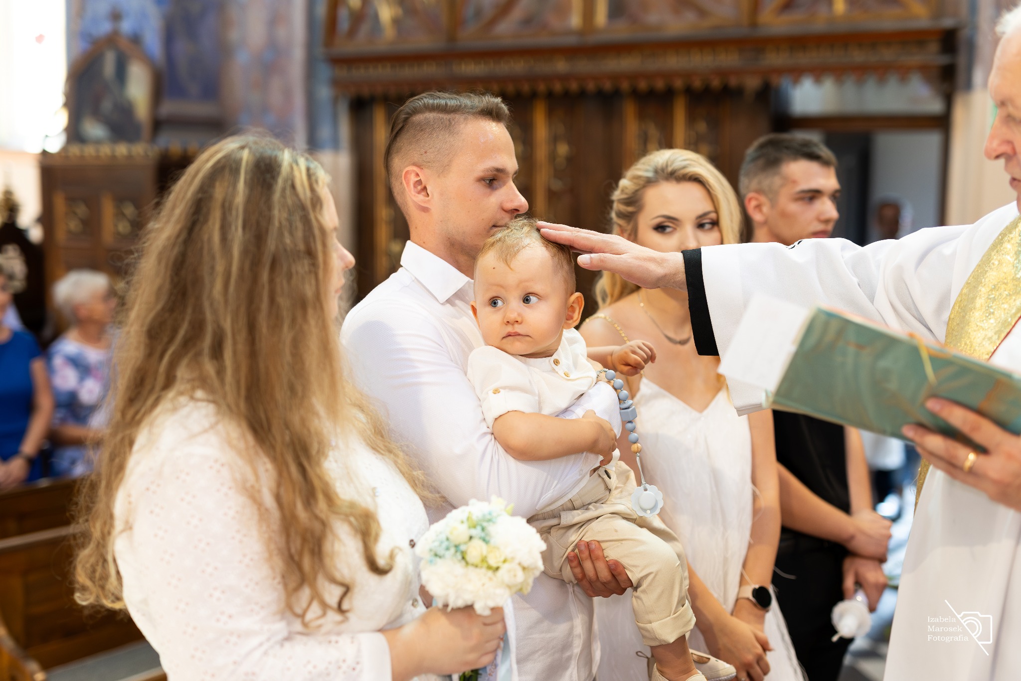 Ksiądz w białej szacie błogosławi niemowlę trzymane przez ojca podczas ceremonii w kościele, obok stoi matka z bukietem białych kwiatów.