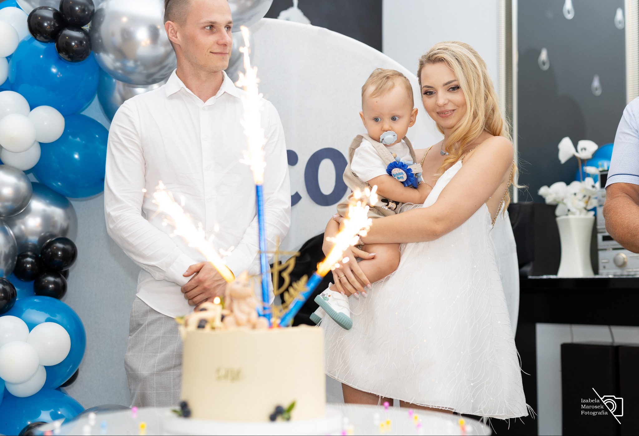 Rodzina świętuje chrzest dziecka z tortem ozdobionym racami, w tle balony w kolorach niebieskim, czarnym i srebrnym.