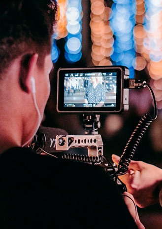 Operator kamery z monitorem nagrywający osobę w tle, widoczne bokeh z oświetlenia ulicznego.