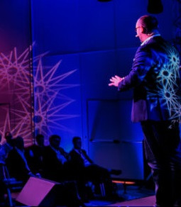 Prelegent w garniturze na scenie, oświetlony niebieskim i różowym światłem, z wyświetlanymi wzorami na ścianie i jego plecach, publiczność siedząca w zaciemnieniu.