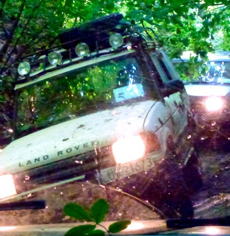 Biały Land Rover z dodatkowym oświetleniem na dachu, w błocie na leśnej trasie off-roadowej, widoczne zielone liście i fragment drugiego pojazdu.